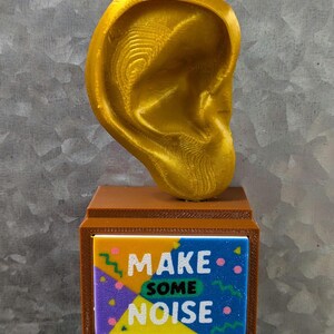 May include: A gold-colored ear sculpture atop a brown rectangular base. The base features a colorful panel with the words "MAKE SOME NOISE" in white, blue, yellow, and purple. The ear is detailed with visible ridges.