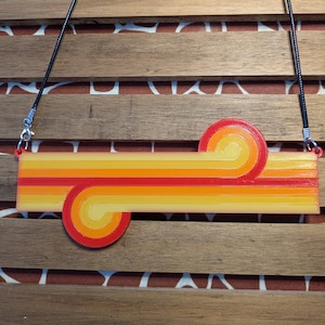 May include: A retro-style necklace with a colorful, layered, abstract design in shades of orange, yellow, and red. The necklace is made of plastic and has a black cord.