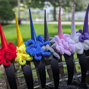 May include: A row of colorful unicorn headbands. Each headband features a black band with a tall, pointed horn and a fluffy, textured mane in various colors, including red, yellow, blue, gray, pink, white, and purple.
