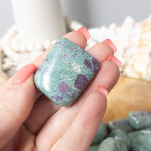 May include: A polished tumbled stone featuring a light green base color with distinct dark magenta and deep red inclusions throughout its surface. The stone has a smooth, rounded rectangular shape with a glossy finish that reflects light.