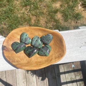May include: A wooden bowl filled with six green and black heart-shaped stones. The stones are arranged in a circle in the bowl.