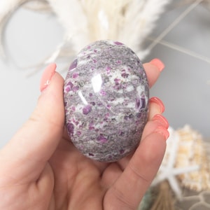 May include: A polished, oval-shaped stone featuring a mottled texture with deep magenta, grey, and white mineral inclusions throughout its smooth, rounded surface. The stone displays a natural, variegated pattern of dark and light mineral deposits.