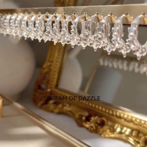 Pode incluir: Brincos de argola transparentes com design de folhas, pendurados em uma barra dourada. Um espelho com moldura dourada está no fundo. O texto "DREAM OF DAZZLE" é visível.