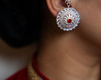 Op de afbeelding: Zilveren oorbel met een cirkelvormig ontwerp en een kleine rode steen in het midden. De oorbel is bevestigd aan een ketting die over het oor gaat. De persoon draagt een rood kledingstuk.
