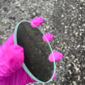 May include: A clear glass circle, approximately 3 inches in diameter, is held in a pink latex glove. The glass is slightly curved and has a smooth, polished edge.