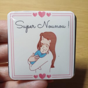 May include: A rectangular white badge with a pink border and the words "Super Nounou!" in cursive. The image features a mother holding a baby, with pink hearts at the top and bottom.