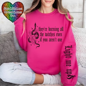 Op de afbeelding: Roze sweatshirt met een zwarte slang en rozen afbeelding en de tekst "they're burning all the witches even if you aren't one". De linkermouw heeft de tekst "Light me ups" in zwart.
