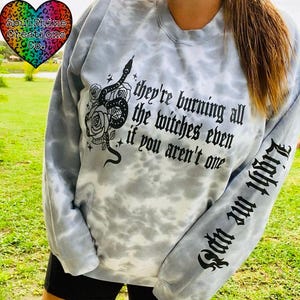 以下が含まれることがあります： 黒の蛇とバラのグラフィックと「they're burning all the witches even if you aren't one」というテキストが書かれたライトブルーのタイダイのトレーナー。右袖には黒で「Light me up」というテキストが書かれている。
