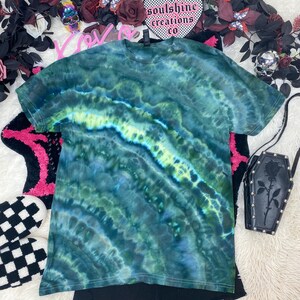 May include: Green, blue, and yellow tie-dye t-shirt with a white trim. The shirt is laid flat on a white surface. The text "soulshine creations co" is visible on a heart-shaped patch.