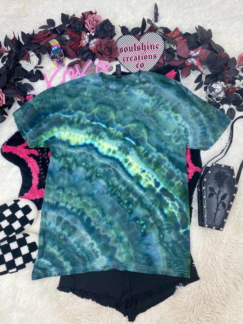 May include: Green, blue, and yellow tie-dye t-shirt with a white trim. The shirt is laid flat on a white surface. The text "soulshine creations co" is visible on a heart-shaped patch.