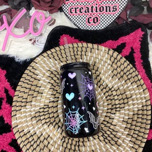 May include: Black tumbler with a spiderweb and bat design. The tumbler has a clear straw and is sitting on a woven placemat. The text "soulshine creations co" is visible on a heart-shaped patch in the background.