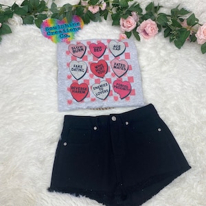 Puede incluir: Una camiseta gris con un patrón a cuadros y corazones rosas con texto rojo. El texto en los corazones incluye frases como "Slow Burn", "One Bed", "Age Gap", "Fake Dating", "Who Hurt You", "Fated Mates", "Reverse Harem", "Enemies to Lovers", y "Forced Proximity". La camiseta se combina con pantalones cortos de mezclilla negros.