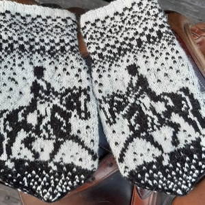 May include: A pair of black and white knitted wool leg warmers with a pattern of riders on horses. The leg warmers are on a brown leather saddle.
