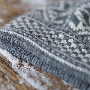 K&ouml;nnte beinhalten: Nahaufnahme einer grauen und wei&szlig;en Strickm&uuml;tze mit traditionellem Muster. Die M&uuml;tze ist aus weichem, flauschigem Garn gefertigt und hat eine strukturierte Oberfl&auml;che.