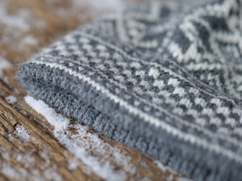 K&ouml;nnte beinhalten: Nahaufnahme einer grauen und wei&szlig;en Strickm&uuml;tze mit traditionellem Muster. Die M&uuml;tze ist aus weichem, flauschigem Garn gefertigt und hat eine strukturierte Oberfl&auml;che.