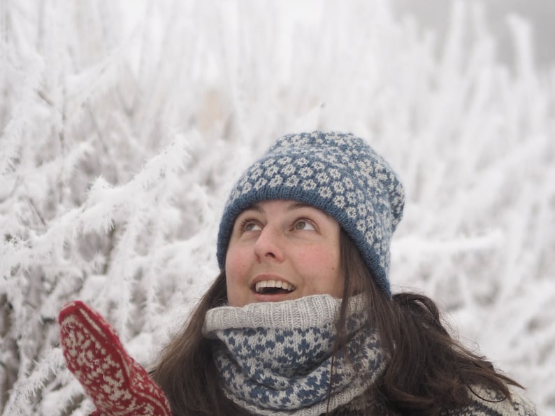 May include: A woman wearing a blue and white knitted hat and matching scarf, looking up at the sky. The background is a snowy, wintery scene.