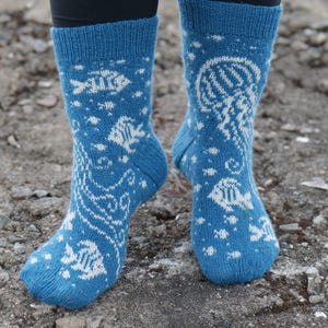 May include: A pair of blue knit socks featuring a white pattern of various fish, a jellyfish, and small bubbles. The socks have a ribbed cuff at the top and a consistent marine-themed design covering the entire surface of the fabric.
