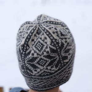 K&ouml;nnte beinhalten: Eine graue und wei&szlig;e Strickm&uuml;tze mit geometrischem Muster. Die M&uuml;tze ist aus Wolle und hat eine weiche, warme Textur.