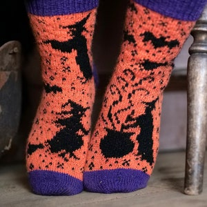 May include: A pair of orange and purple socks with a black witch and cauldron pattern. The socks are knitted and have a festive Halloween theme.