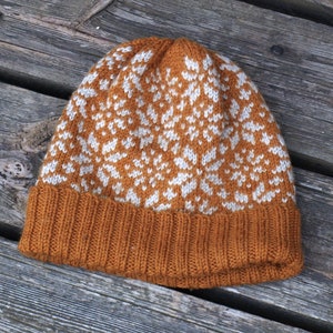 Può includere: Un cappello di lana lavorato a maglia marrone e bianco con un motivo a fiocco di neve. Il cappello è in lana e ha un bordo ripiegato.