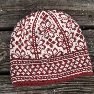 Puede incluir: Un gorro de punto rojo y blanco con un patrón geométrico. El gorro está hecho de lana y tiene una sensación cálida y acogedora.