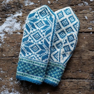 Pode incluir: Um par de luvas de malha azul e branca com um padrão geométrico. As luvas estão sobre uma superfície de madeira com neve.