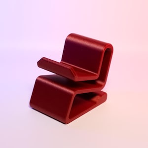May include: A modern, minimalist phone stand in a deep red color. The stand has a unique, curved design with a small ledge to hold a phone. The stand is made of a solid material and is suitable for a desk or table.