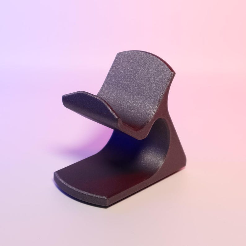 Pode incluir: Um suporte de telefone cinza escuro com uma base curva e um ber&ccedil;o para segurar um telefone. O suporte tem uma superf&iacute;cie texturizada e um design minimalista. O fundo tem um gradiente de rosa e roxo.