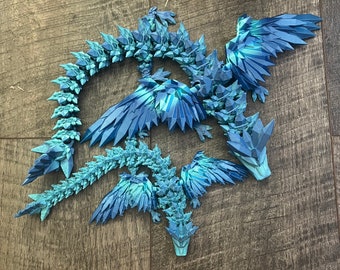 May include: A detailed, blue and teal dragon sculpture with a segmented body, wings, and a pointed head. The dragon is made of geometric shapes, giving it a crystalline appearance. The sculpture is lying on a wooden surface.