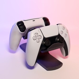 Op de afbeelding: Twee witte en zwarte gamecontrollers, waarvan één op een zwarte standaard rust. De controllers hebben een strak ontwerp met diverse knoppen en directionele pads. De achtergrond heeft een verloop van roze en paars.
