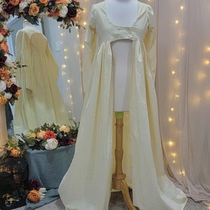 May include: A pale yellow Regency-style dress on a mannequin. The dress features long sleeves, a high waist, and a flowing skirt. The fabric has a textured appearance. The background includes a mirror reflecting the dress, floral arrangements, and string lights. The text "Being Sew Weird" is visible.