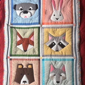 Könnte beinhalten: Eine handgefertigte Steppdecke mit sechs Tiergesichtern: einem Waschbären, einem Kaninchen, einem Fuchs, einem Bären, einem Wolf und einem Otter. Jedes Tier ist in einem Quadrat mit einem anderen farbigen Hintergrund angeordnet. Die Steppdecke hat einen weißen Rand mit einem Streifenmuster.
