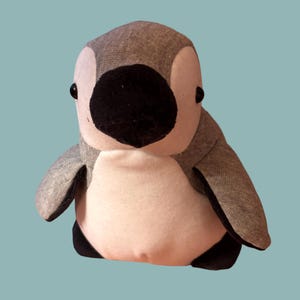 Könnte beinhalten: Ein Plüsch-Pinguin mit einem Kopf und Rücken in Grau meliert, einem cremefarbenen Bauch und einem einfarbig schwarzen Schnabel. Das Spielzeug hat kleine schwarze Knopfaugen und schwarze Flossen sowie eine abgerundete Form aus weichem Stoff.