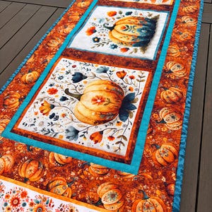 Könnte beinhalten: Ein rechteckiger Tischläufer aus Stoff mit einem sich wiederholenden Muster aus orangefarbenen Kürbissen und Blumenmotiven auf weißem Grund, umrahmt von Bordüren mit dunkelorangen Kürbisdrucken und leuchtend türkisfarbenen Stoffstreifen.