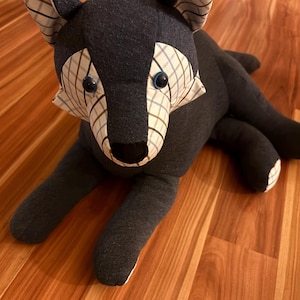 Könnte beinhalten: Ein Stofftier in Form eines Wolfes, gefertigt aus dunkelgrauem Stoff und weißem Stoff mit blau-braunem Karomuster. Der Wolf hat zwei dunkelblaue Knopfaugen, eine schwarze Stoffnase und spitze Ohren mit karierten Akzenten.