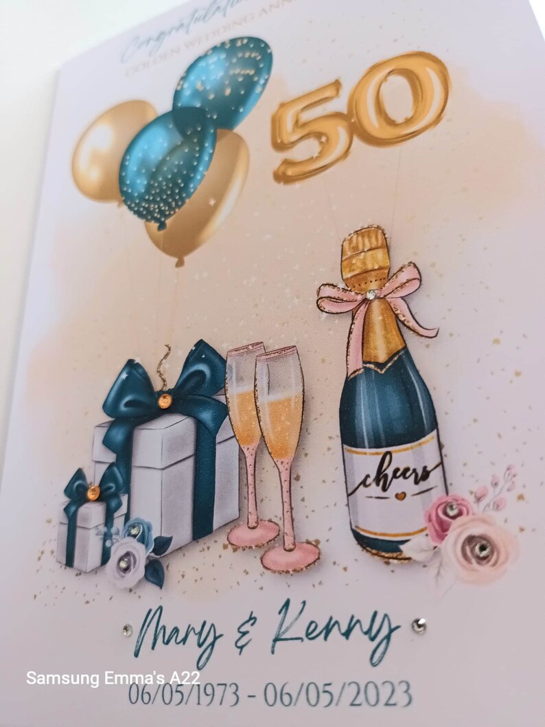 May include: A gold 50th anniversary card with a champagne bottle, two champagne flutes, and two gift boxes with teal and white ribbons. The card has a pink and white floral design with gold glitter. The text on the card reads "Mary & Kenny" and "06/05/1973 - 06/05/2023".