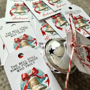 May include: A collection of white rectangular gift tags featuring a gold bell illustration with a red ribbon and the text THE BELL STILL RINGS FOR THOSE WHO TRULY BELIEVE, accompanied by a large silver jingle bell tied with red and white twine.