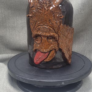 Pode incluir: Um vaso de cerâmica marrom com um rosto esculpido e uma língua vermelha para fora. O vaso está em uma plataforma giratória preta.