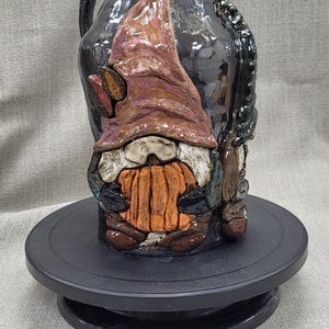 May include: A black ceramic pitcher with a gnome holding a pumpkin. The gnome is wearing a purple hat and has a white beard. The pitcher has a handle and is decorated with green leaves.