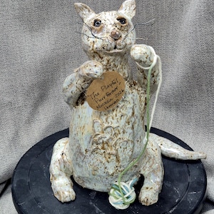 May include: A ceramic cat figurine with a playful pose, holding a ball of yarn. The cat is off-white with brown accents and has a tag that reads "The Playful Purrformer". The figurine is on a black surface.