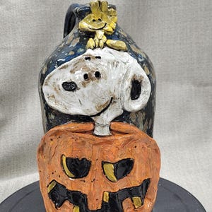 Puede incluir: Una jarra de cerámica decorada con una calabaza, un perro y un pájaro. La jarra está pintada en tonos de azul, blanco, naranja y amarillo.