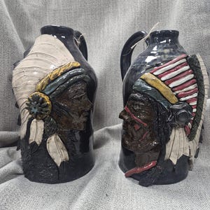 May include: Two ceramic jugs with dark blue and black glaze. Each jug features a relief of a Native American profile with a feathered headdress. One jug has a white and tan design, the other has red and white stripes.