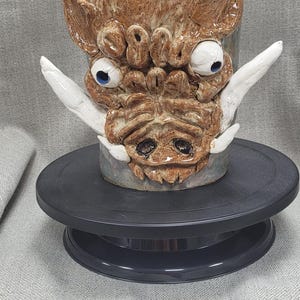 Könnte beinhalten: Eine Keramikvase in Form eines Eberkopfes mit weißen Hauern und blauen Augen. Die Vase hat eine braune und weiße Glasur und steht auf einer schwarzen Töpferscheibe.