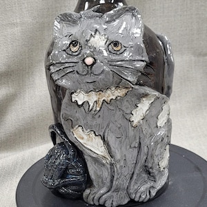 May include: A ceramic cat figurine with gray and white fur, sitting on a black base. The cat is holding a ball of yarn in its paws.