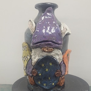 May include: A ceramic gnome vase with a purple hat, holding a blue and green patterned disc. The vase is decorated with colorful patterns and has a textured surface.