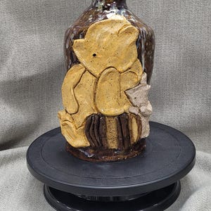 Op de afbeelding: Een handgemaakte keramische vaas met een Winnie de Poeh-ontwerp. De vaas is voornamelijk bruin met een gele berenfiguur en wordt gepresenteerd op een zwarte draaiende standaard. Een uniek decoratief item.