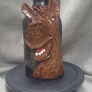 May include: A ceramic vase with a brown donkey face sculpted on the front. The vase is glazed in a dark brown color and has a glossy finish.