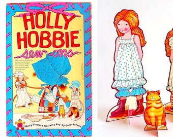 May include: A Holly Hobbie Sew Ons activity toy set by Colorforms featuring a rectangular box with blue borders and pink ribbon illustrations, alongside two paper doll figures with red hair and floral dresses and a small orange cat figure.