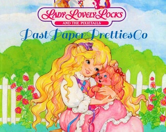 Peut inclure: Une couverture de livre à colorier représentant Lady Lovely Locks et les Pixietales. La couverture montre une fille aux longs cheveux blonds tenant un chiot rose. Le texte "Past Paper Pretties Co" est en lettres bleues.