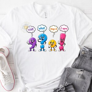 Może przedstawiać: Biały t-shirt z grafiką przedstawiającą cztery antropomorficzne znaki interpunkcyjne: fioletowy przecinek, niebieski znak zapytania, żółte koło i różowy wykrzyknik. Każda postać ma dymek z tekstem wait, what, stop it oraz I agree.
